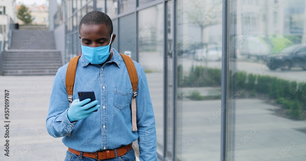 Obraz premium Close up of African American young man in medical mask walking the street and talking on mobile phone. Male pedestrian in respiratory protection strolling and speaking on cellphone. Conversation.