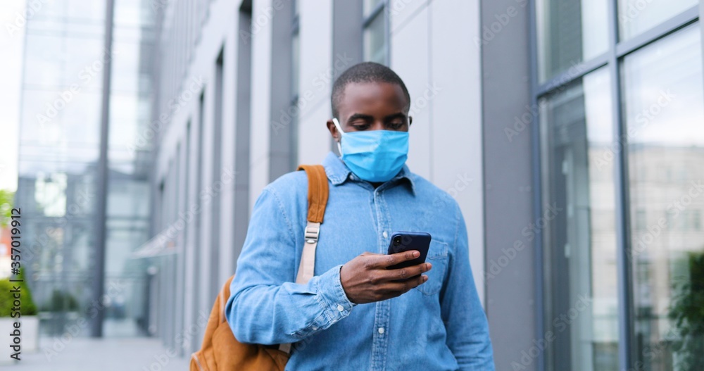 African American man in medical mask standing on street and texting message on mobile phone. Male in respiratory protection is outdoors and tapping or scrolling on smartphone. Browsing online. African American man in medical mask standing on street and texting message on mobile phone. Male in respiratory protection is outdoors and tapping or scrolling on smartphone. Browsing online.