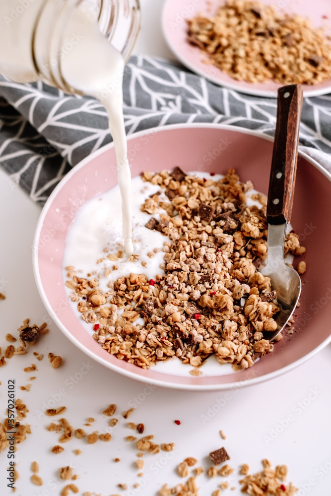 Granola muesli with fresh milk and pieces of chocolate in a pink plate on a white table. Healthly food, breakfast cooking concept. Granola muesli with fresh milk and pieces of chocolate in a pink plate on a white table. Healthly food, breakfast cooking concept.