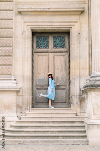 Charming elegant young woman wearing blue dress, posing in front of ancient wooden door of old vintage building with one leg up