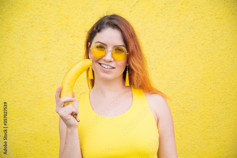 Portrait of a beautiful red-haired woman in a yellow dress and sunglasses holds a banana near sensual lips. Girl posing with fruit on a yellow background. Portrait of a beautiful red-haired woman in a yellow dress and sunglasses holds a banana near sensual lips. Girl posing with fruit on a yellow background.