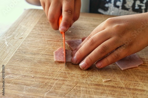 Little boy helping out in the kitchen by cutting ham with an orange knife on a wooden cutting board