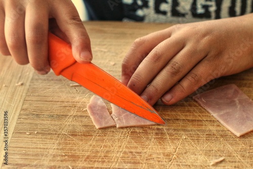 Little boy helping out in the kitchen by cutting ham with an orange knife on a wooden cutting board