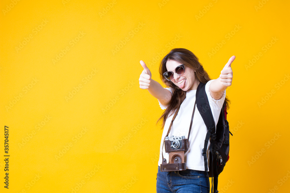 Cool travel tourist girl showing thumb up approval like gesture,satisfied recommend.Excited young woman in white t-shirt,backpack,sunglasses,retro photo camera isolated on yellow background.copy space Cool travel tourist girl showing thumb up approval like gesture,satisfied recommend.Excited young woman in white t-shirt,backpack,sunglasses,retro photo camera isolated on yellow background.copy space