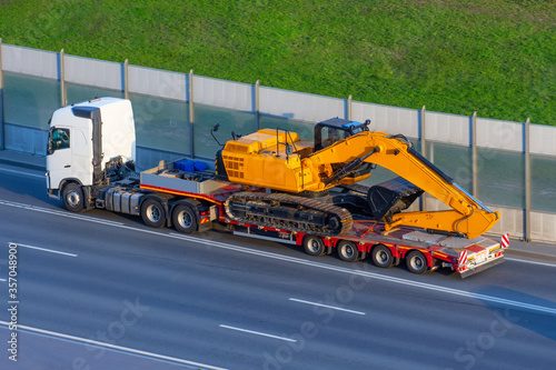 Heavy new yellow excavator on transportation truck with long trailer platform on the highway in the city.