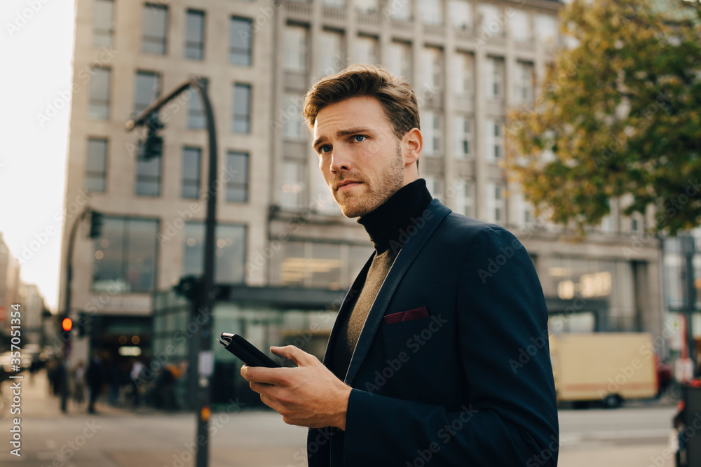 Contemplating businessman with smart phone looking away while standing against building in city Contemplating businessman with smart phone looking away while standing against building in city