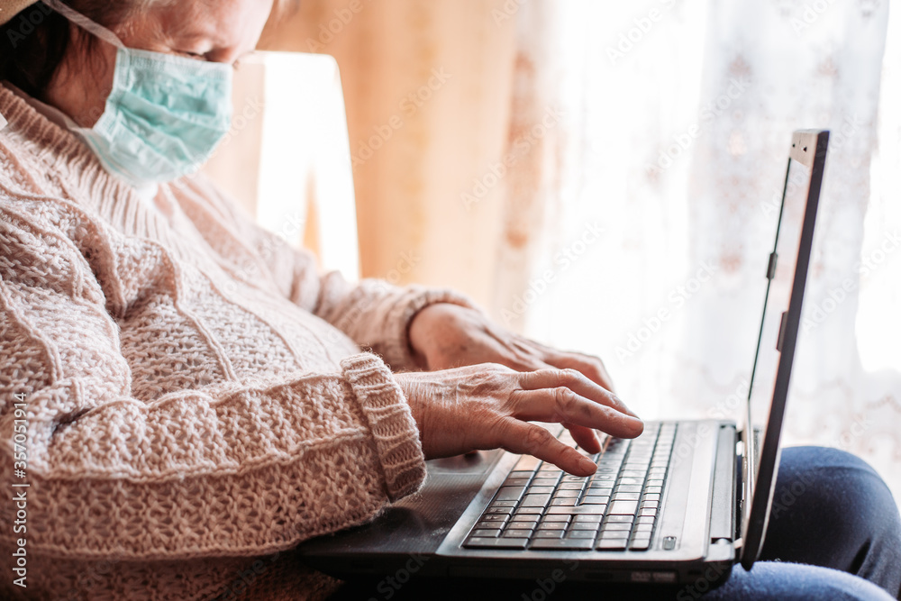 Fototapeta premium Elderly woman wearing a medical mask using her laptop, typing something.
