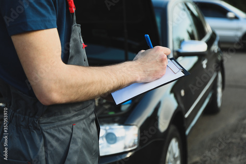 A man in overalls writes to a clipboard. Mechanic. Car inspection photo