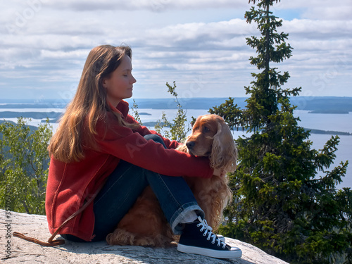A young woman sitting on a stone plateau and with tenderness and love looking at the English Cocker Spaniel red color. In the background you can see spruce, shrubs and a large lake with Islands.