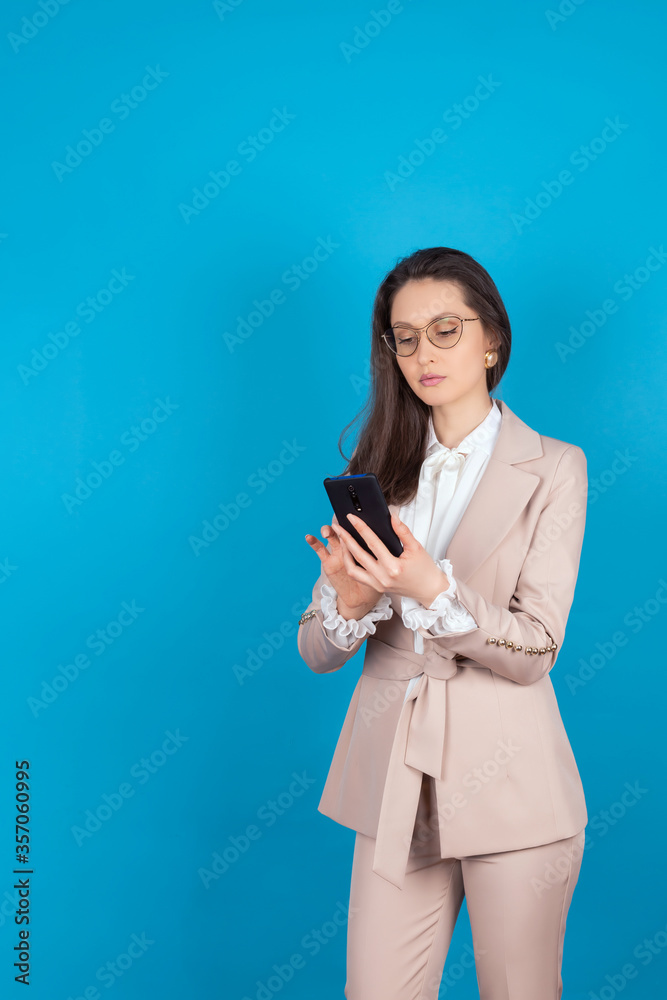 Portrait of a beautiful confident business girl in glasses and a light suit, with a phone in her hands. A young woman makes purchases online. Isolate on a blue background. Copy space. Mock up Portrait of a beautiful confident business girl in glasses and a light suit, with a phone in her hands. A young woman makes purchases online. Isolate on a blue background. Copy space. Mock up