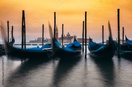 Gondolas docked at Grand Canal in Venice with Church in background. Motion blur of moving gondolas in water of canal.