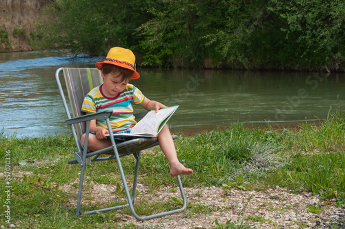 Cute little boy of 4 years old in an orange hat and a striped T-shirt is sitting on a folding chair with a book on the green lawn by the river. Summer, vacations, happy childhood in nature.