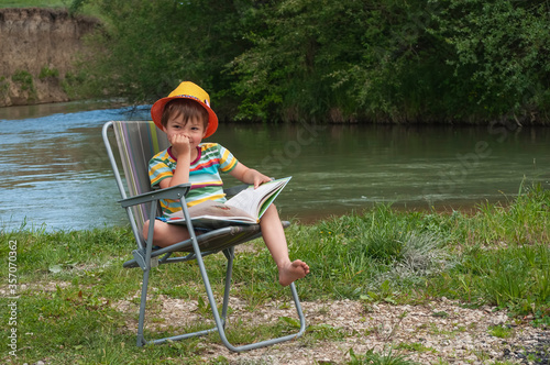 Cute little boy of 4 years old in an orange hat and a striped T-shirt is sitting on a folding chair with a book on the green lawn by the river. Summer, vacations, happy childhood in nature.