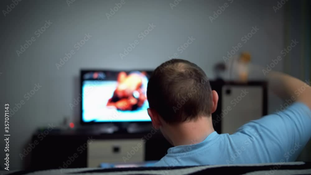 Young energetic man watches a sports game on TV at home, sits on the sofa in the evening, strong emotions Young energetic man watches a sports game on TV at home, sits on the sofa in the evening, strong emotions