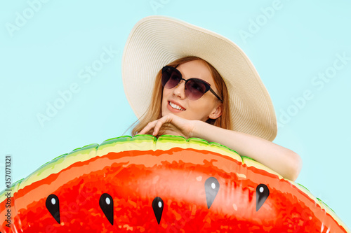 pensive beautiful girl in a summer hat and sunglasses  dressed in a swimsuit  stands with an inflatable ring on an blue background