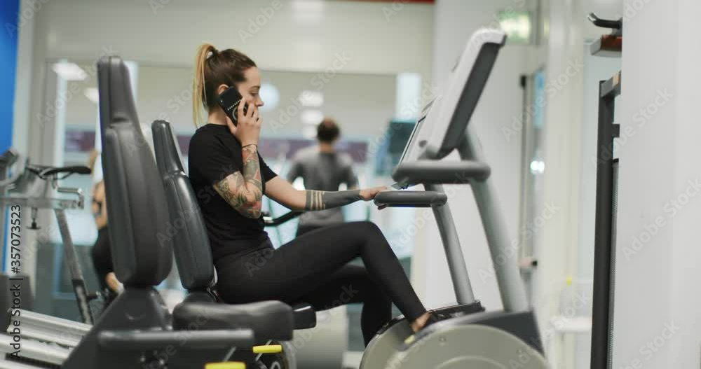 woman exercising with elliptical cross trainer while talking on cellphone at gym.Side view, slow motion. Woman training at elliptical bike while talking on mobile phone.Woman pedaling using smartphone woman exercising with elliptical cross trainer while talking on cellphone at gym.Side view, slow motion. Woman training at elliptical bike while talking on mobile phone.Woman pedaling using smartphone