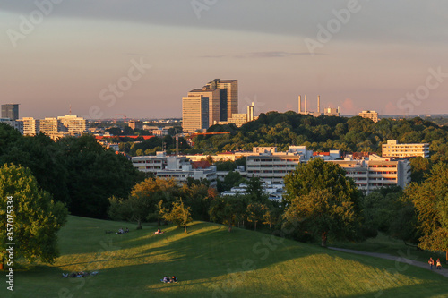 People relaxing during sunset time on an artificial hill amidst urban greenery with modern urban skyscrapers in the background in the German city of Munich