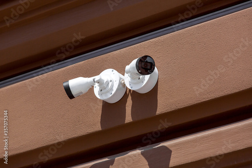 two white CCTV cameras installed on the exterior facade of a brown wall, close up security surveillance systems and remote order control on sunny day.