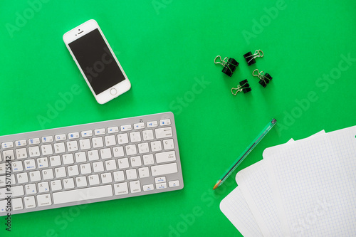 Mobile phone, wireless keyboard, pen, papers and clips on green background, office concept