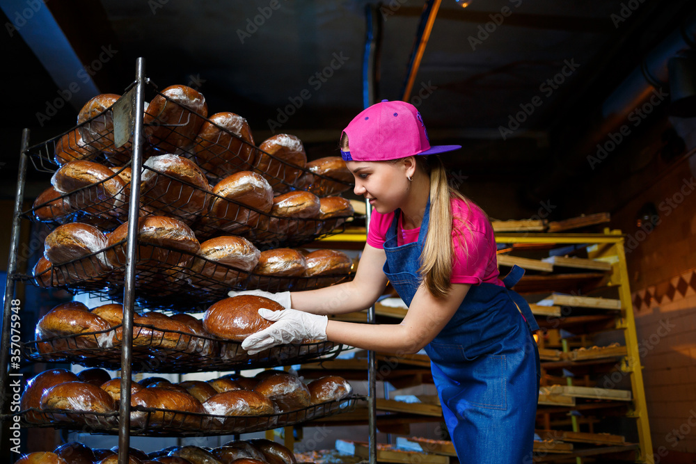 A young girl works in a bakery. She puts bread on a shelf. Woman baker at workplace in a bakery. A professional baker holds bread in his hands. Bread production concept