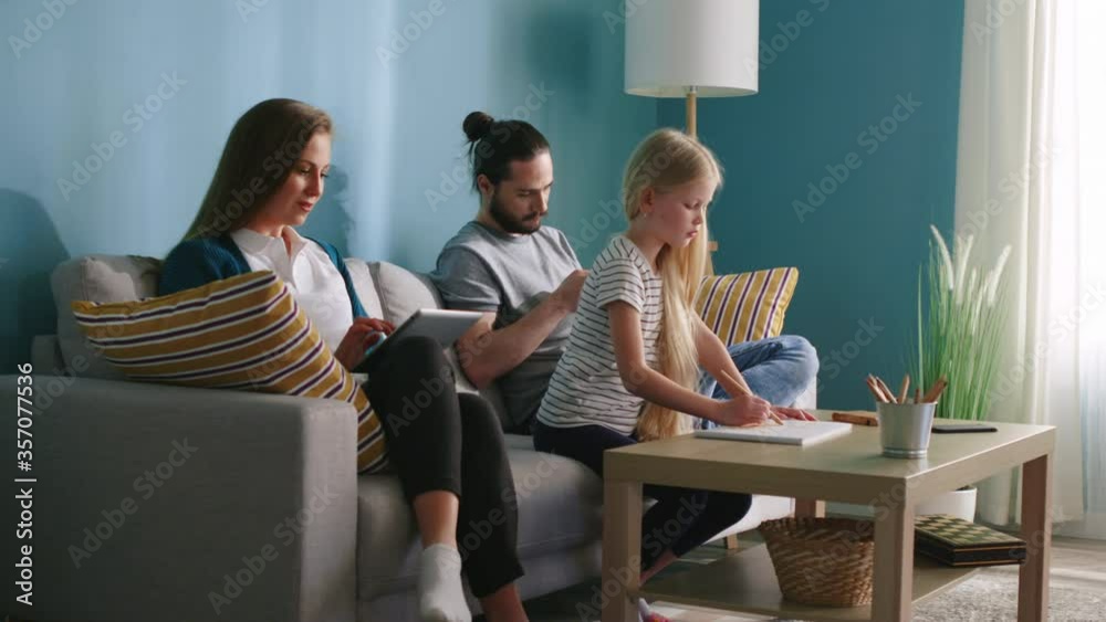 Young family prefers indoor leisure, spending time in cozy living room, but everyone is busy with own hobby - mother is shopping online, father looks for news in social media, and daughter is drawing. Young family prefers indoor leisure, spending time in cozy living room, but everyone is busy with own hobby - mother is shopping online, father looks for news in social media, and daughter is drawing.
