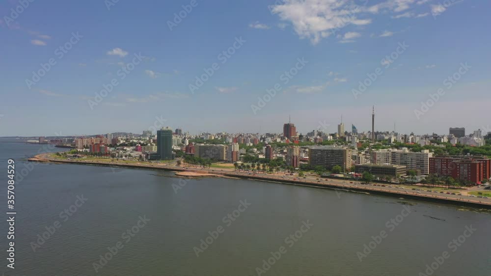 Aerial shot of vehicles on street by buildings in city, drone flying forward towards coastline against sky on sunny day - Montevideo, Uruguay Aerial shot of vehicles on street by buildings in city, drone flying forward towards coastline against sky on sunny day - Montevideo, Uruguay