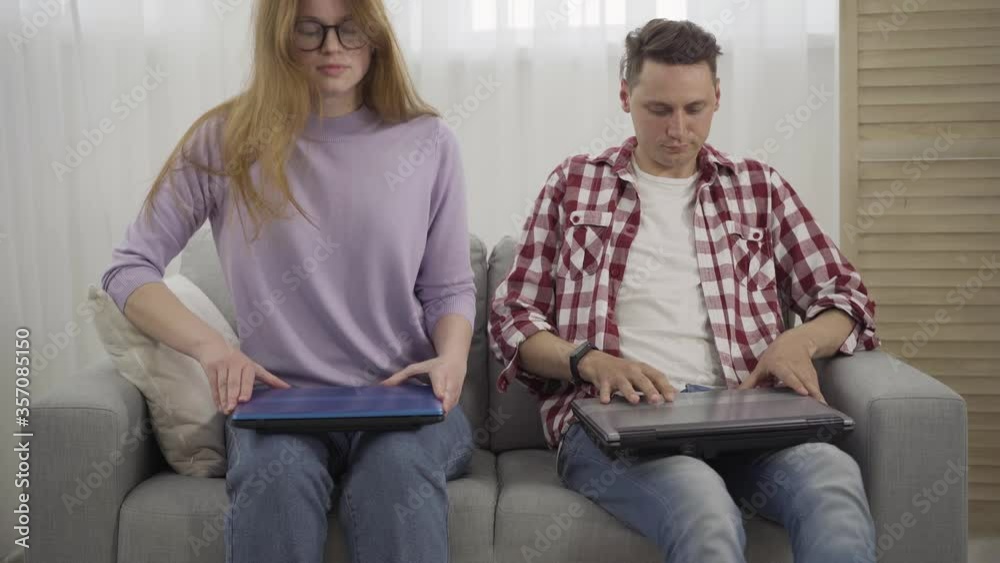 Confident Caucasian redhead woman and brunette man sitting down on couch and opening laptops. Portrait of serious young male and female freelancers working online. Freelance, lifestyle. Confident Caucasian redhead woman and brunette man sitting down on couch and opening laptops. Portrait of serious young male and female freelancers working online. Freelance, lifestyle.