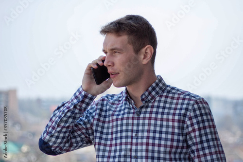 Caucasian  men  is making a business call. There is a city on the back ground.  Dressed into checkered shirt. Stands on the 9th floor. photo