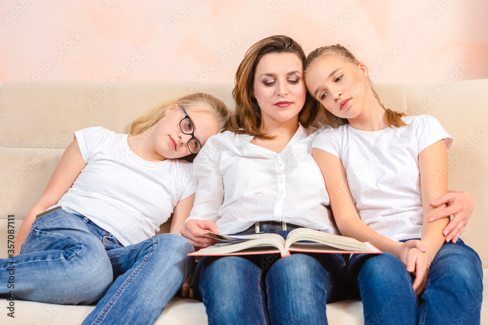 Portrait of Young Caucasian Mother Reading a Book Together with Her Twin Daughters While Sitting on Sofa Indoors. Portrait of Young Caucasian Mother Reading a Book Together with Her Twin Daughters While Sitting on Sofa Indoors.