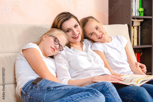 Portrait of Young Happy Caucasian Mother Reading a Book Together with Her Twin Daughters While Sitting on Sofa Indoors.