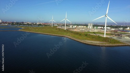 Aerial panning shot of wind turbines in city by sea against blue sky, drone flying over coastline on sunny day - Copenhagen, Denmark