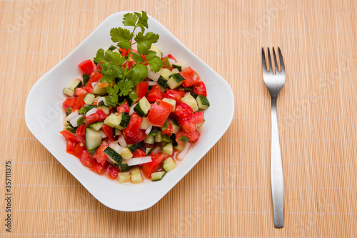 Fresh salad of finely chopped cucumbers, tomatoes and onions with sunflower oil in a white plate and a fork on a wooden table. The concept of healthy nutrition and eco-friendly, vegetarian diet.