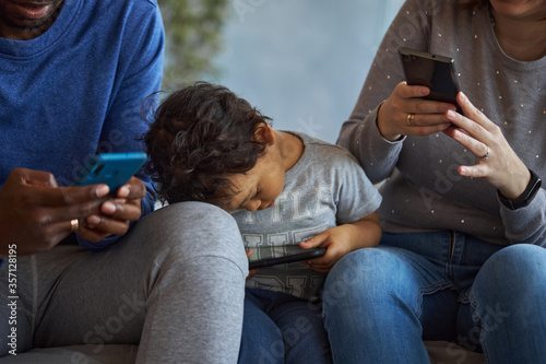 Concentrated members of family sit together on one sofa. Each of them has phone in hands. They look at them.