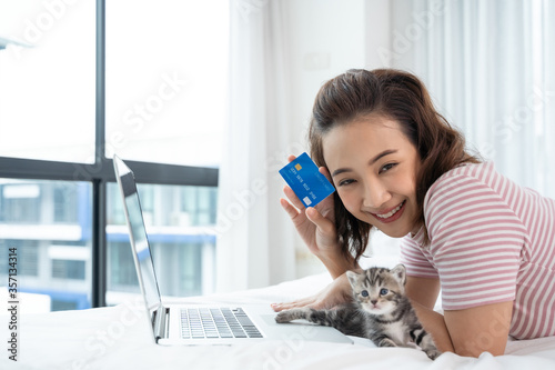 An Asian woman holds a credit card and is happily shopping online in her bed. And there is a cat beside. Concepts online shopping , life style, stay home, banks, financial transactions.