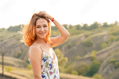 Tender beautiful red-haired girl enjoys the sunset in a field with a hill. © Kate