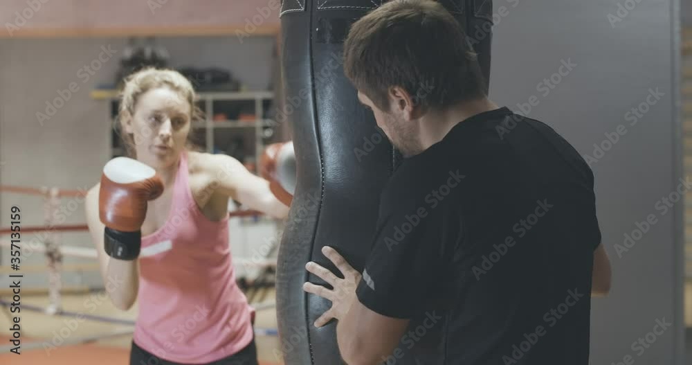 Back view of professional coach holding punching bag and endorsing young blond woman punching equipment. Personal trainer training female self-defense or boxing in gym. Cinema 4k ProRes HQ. Back view of professional coach holding punching bag and endorsing young blond woman punching equipment. Personal trainer training female self-defense or boxing in gym. Cinema 4k ProRes HQ.
