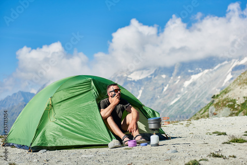 Man tourist sitting inside camp tent and using walkie-talkie. Male traveler with radio in hand resting in tourist tent with mountains and blue sky on background. Concept of travelling, hiking, camping