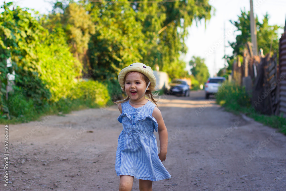Happy little girl in a hat and dress laughs, runs playing on a summer street. Child walks on a village road.
The concept of happy childhood.