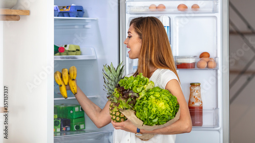 Only the freshest produce for my fridge! A healthy young woman putting a container of salad into the fridge. Woman at home opening the fridge and taking food out while looking at the camera