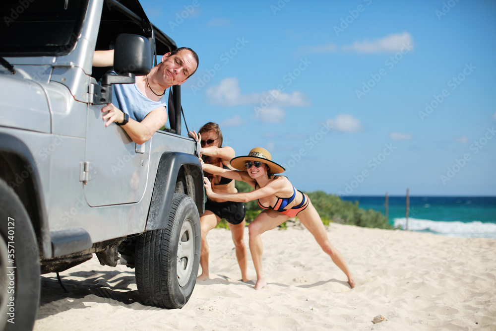 Naklejka premium The car stuck in the sand.Two beauty and sexy women in bikini pushing a off road car near sea on beach while man is emboldening them.Transportation,teamwork,funny concept.