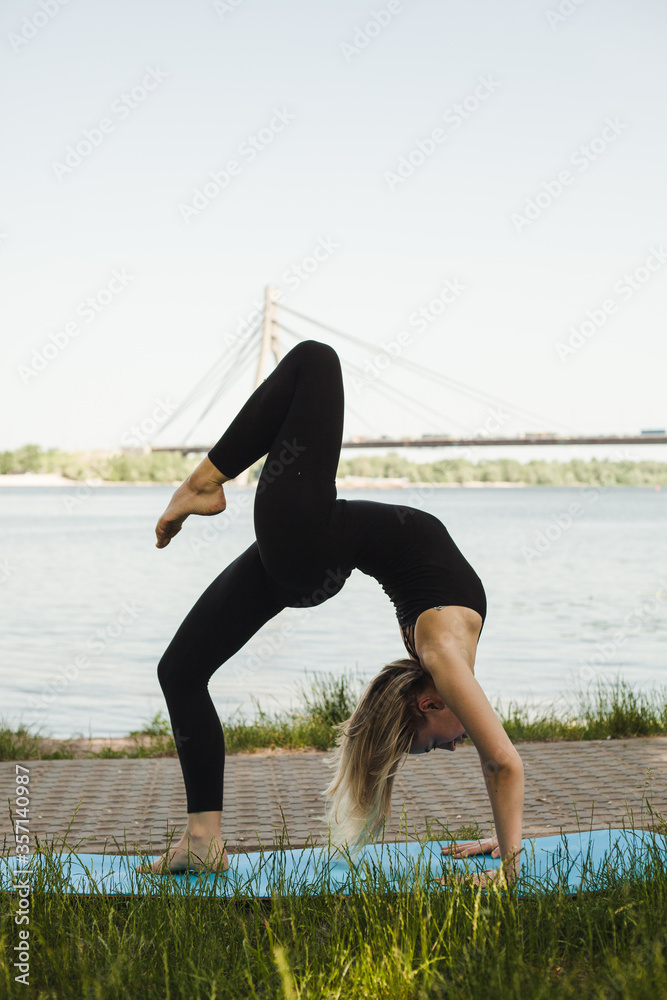 Fototapeta premium Portrait of a gorgeous young woman practicing yoga outdoors in a park. Beautiful girl in a black skinny suit. Calm and relax, feminine happiness. Horizontal, blurred background. 