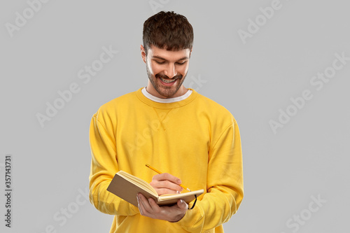 people concept - smiling young man in yellow sweatshirt writing to diary over grey background