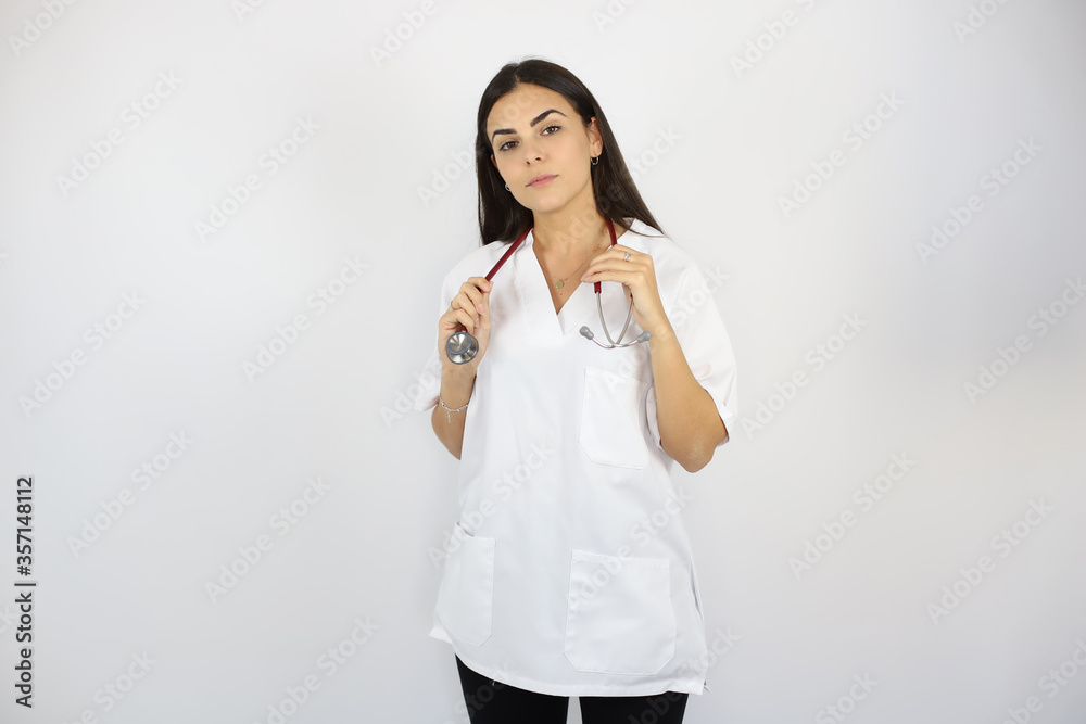 Young beautiful woman standing over white isolated background wearing her doctor uniform smiling and picking up her stethoscope Young beautiful woman standing over white isolated background wearing her doctor uniform smiling and picking up her stethoscope