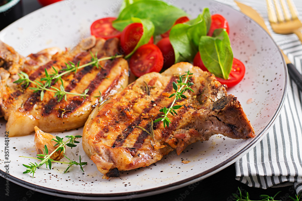 Grilled pork steaks and salad with tomatoes in plate on dark background.