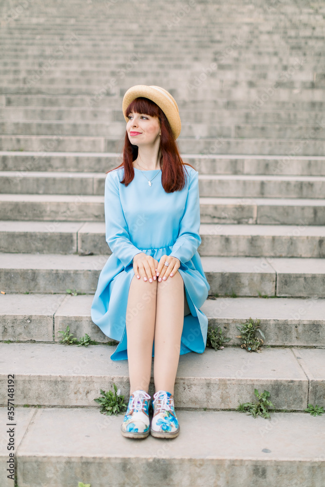 Image of beautiful stylish woman in blue dress sitting on street stairs in old European city.