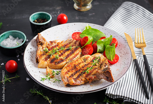 Grilled pork steaks and salad with tomatoes in plate on dark background.