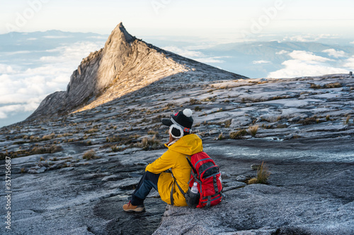 Young woman trekker with backpack sitting in front of South peak of Kinabalu mountain massif, Borneo island in Sabah state, Malaysia