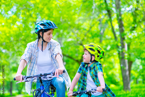 A smiling child in a bicycle helmet looks at his mother in a bicycle helmet while walking great in the park.