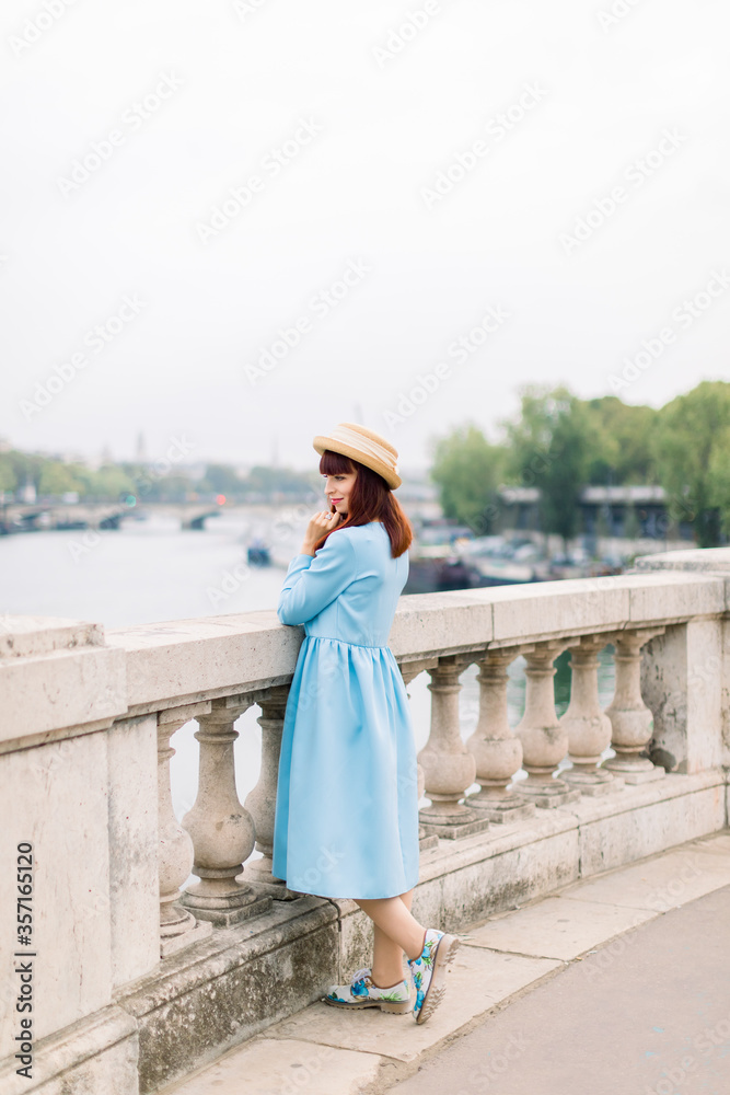 Young attractive woman in blue dress and straw hat, enjoying great view on the Paris and Seine river, standing on the bridge and looking away