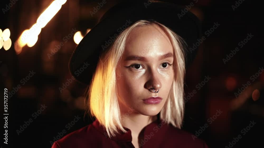 Portrait of trendy hipster woman with blond hairstyle standing on city street at night. Hat, nose piercing. Mysterious girl. Slow motion. Portrait of trendy hipster woman with blond hairstyle standing on city street at night. Hat, nose piercing. Mysterious girl. Slow motion.